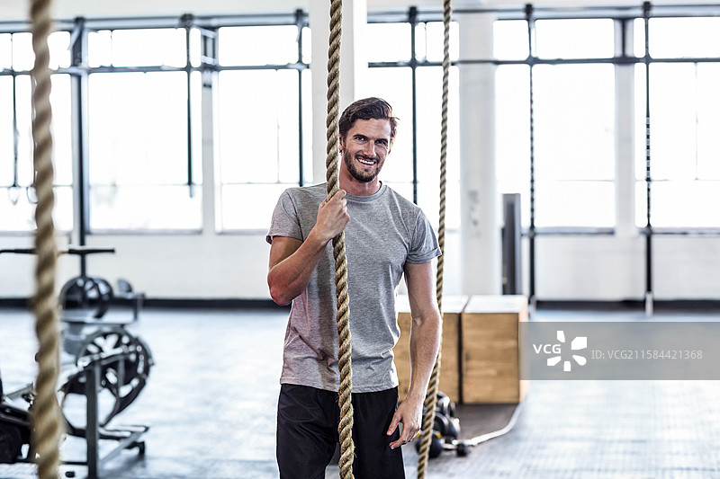 男子在健身房内抓住粗大的攀岩绳，身穿灰色运动T恤和短裤，周围有重量架。图片素材