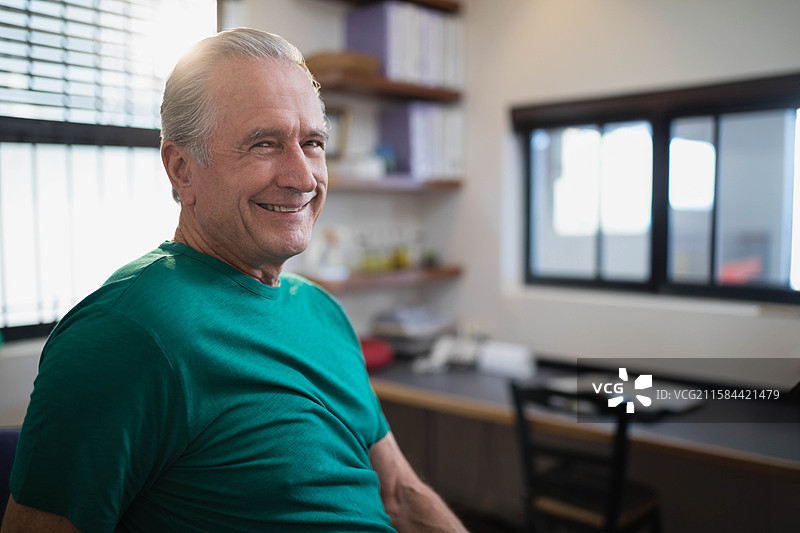 穿着绿色T恤的老年男子坐在书房的桌子旁，桌上有文件和笔记本，留有空白空间图片素材