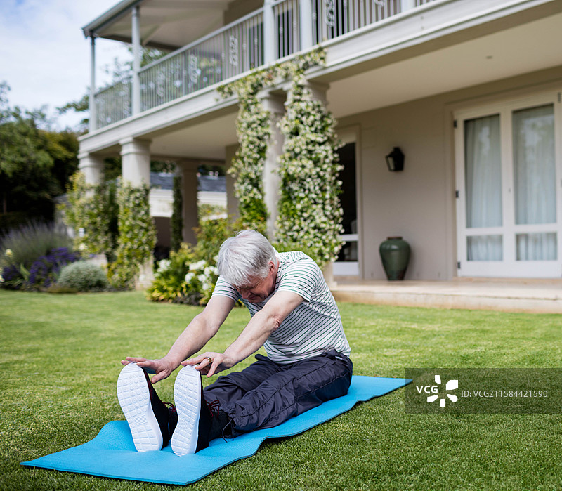 一位年长男性在后院草坪上穿着运动鞋，在蓝色瑜伽垫上进行前屈拉伸。图片素材