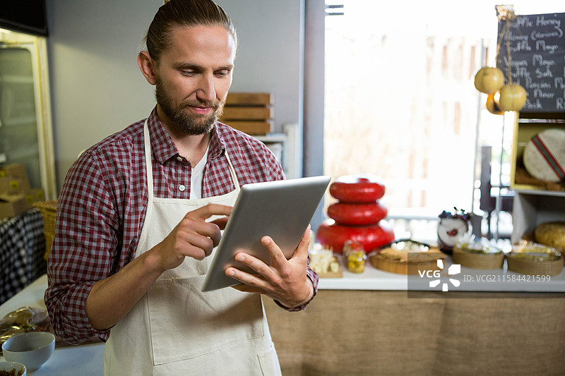 男子在奶酪店的柜台后使用平板电脑，周围是红蜡包裹的奶酪轮和篮子。图片素材