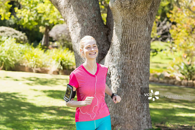 穿着运动服的女性在公园里跑步，经过橡树，手臂上挂着手机臂带，佩戴着健身手表。图片素材