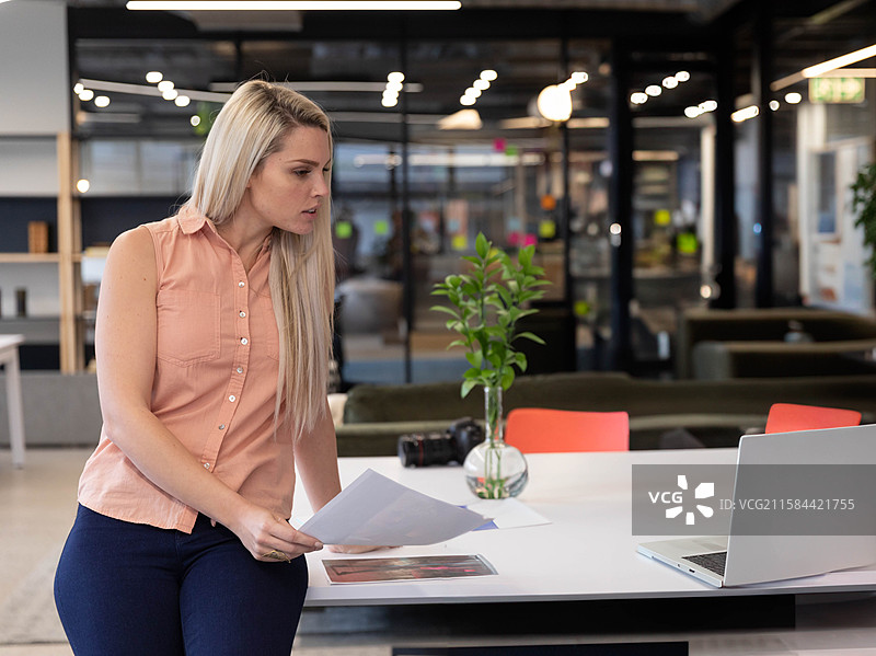 银色笔记本电脑静静地放在现代开放式办公室的桌子上，旁边是单反相机和一个绿叶花瓶。图片素材