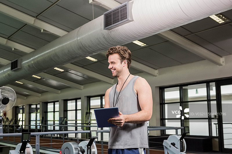 男性教练在健身房里拿着平板电脑查看数据，旁边有划船机、风扇和露出的管道。图片素材