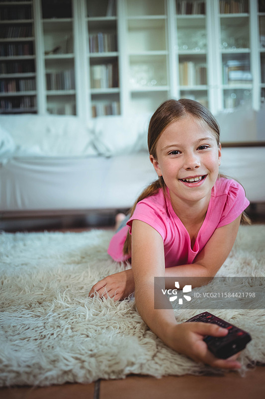 女孩躺在客厅的白色长毛地毯上，伸手去拿沙发旁的遥控器和书架上的书籍。图片素材
