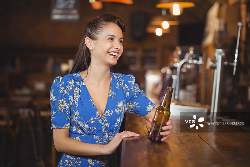 女人坐在木质酒吧柜台前，手里拿着棕色啤酒瓶，微笑着，留有空白空间。图片素材