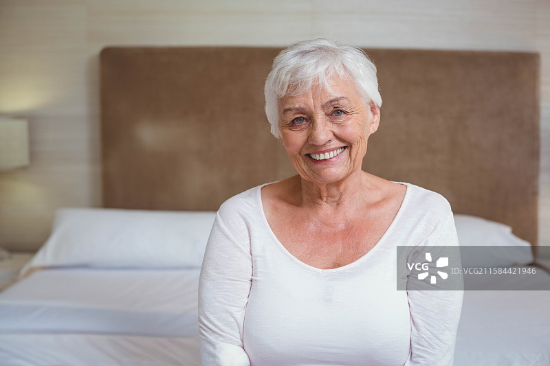 一位老年女性坐在卧室床边，床头有软垫靠背，床头柜上有一盏台灯。图片素材