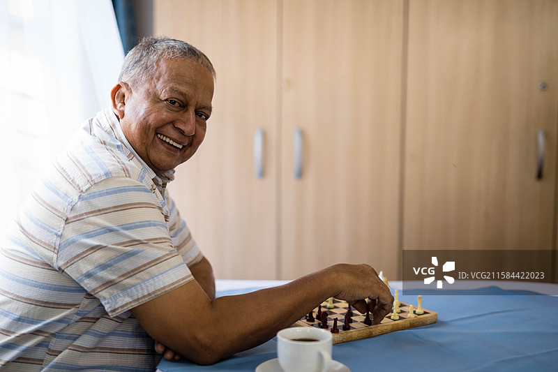 一位微笑的印度老人在家中喝着咖啡，移动棋盘上的棋子。图片素材