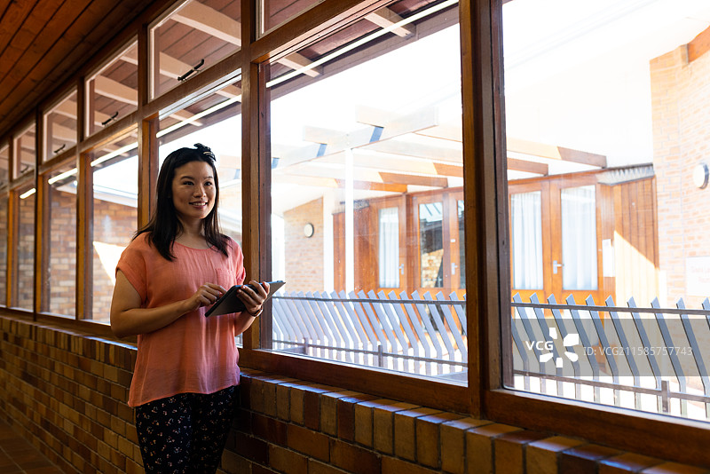 亚洲教师站在阳光明媚的走廊里，手持平板电脑，留有空白空间图片素材