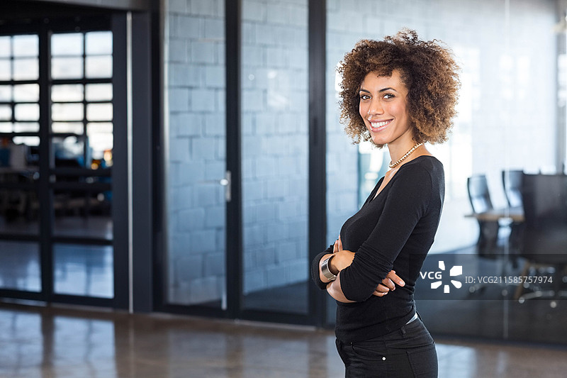 专业女性双臂交叉站在现代办公室，周围有玻璃隔断和办公桌。图片素材