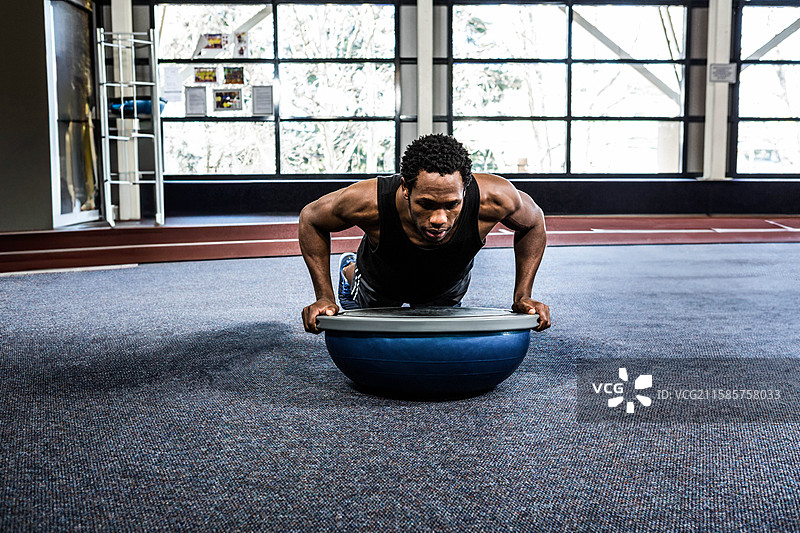 健身训练者在健身房的蓝色BOSU球上做俯卧撑，身后是红色跑道。图片素材