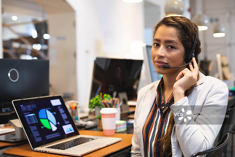穿着西装的女性在办公室使用耳机接听电话，电脑上显示着图表，留有空白空间。图片素材