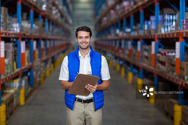仓库工人在过道的货架上检查库存水平，手里拿着夹板和笔。图片素材