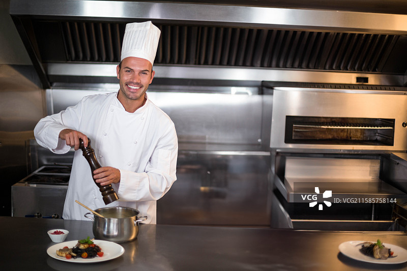 男性厨师在商业厨房的钢制台面上，用木制胡椒磨对锅中的食材进行调味。图片素材