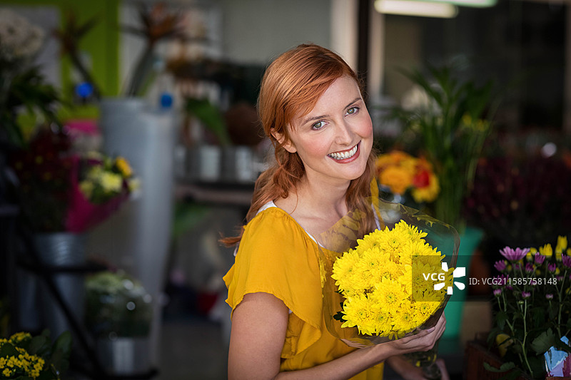 女花店老板手持包裹好的黄色菊花花束，店内有水桶、盆栽植物和花瓶。图片素材