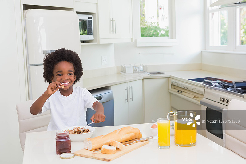 非裔美国男孩坐在白色厨房桌子旁，手里拿着勺子，正在碗里吃麦片。图片素材