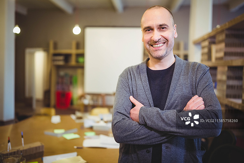 一位四十多岁的男性站在工作空间里，双臂交叉，微笑着，旁边的桌子上散落着便利贴。图片素材