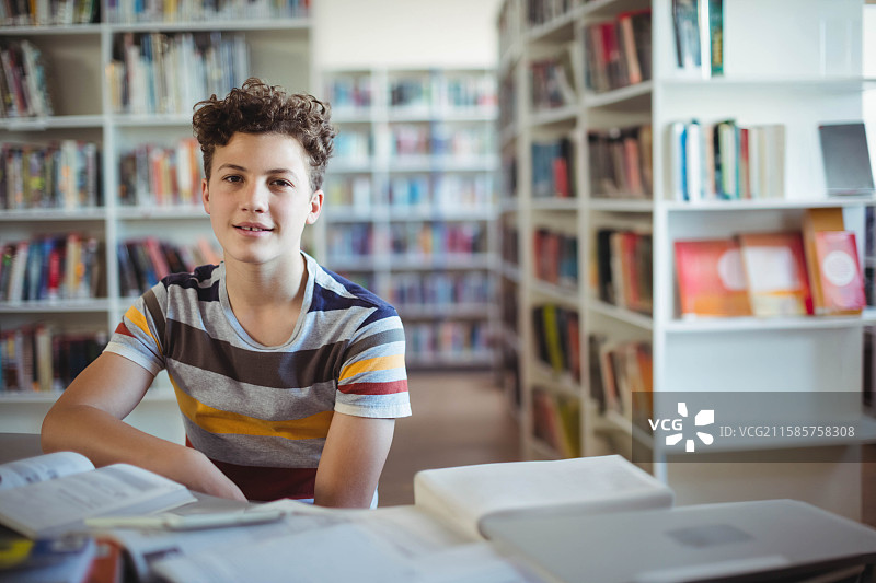 少年男孩坐在图书馆的学习桌前，周围是打开的教科书和笔记本电脑。图片素材