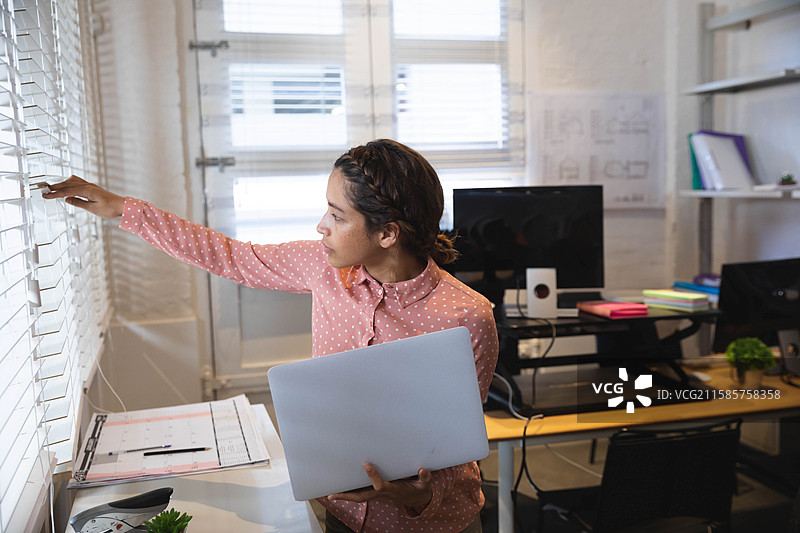 非裔美国女性在设计工作室调整百叶窗，手持笔记本电脑和夹板，留有空白空间图片素材