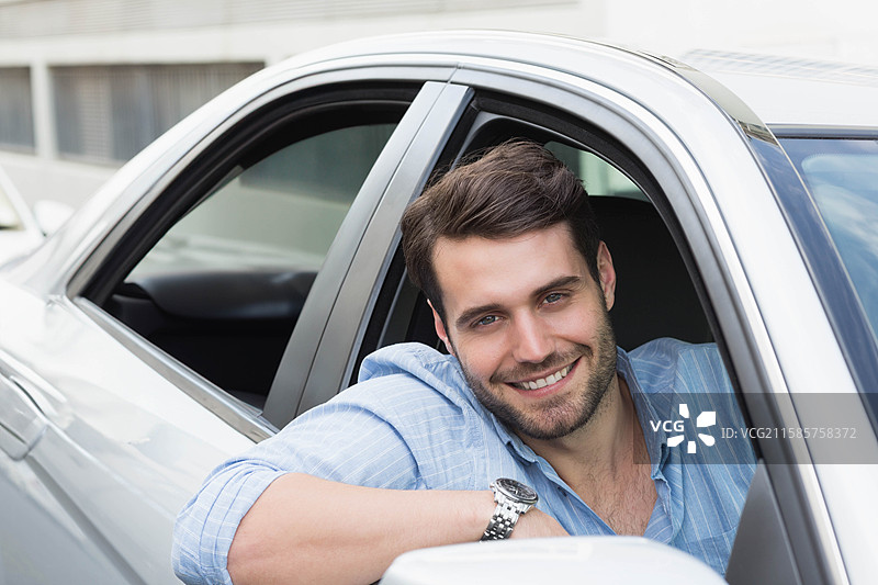 一名成年男性司机微笑着握着银色轿车的方向盘，旁边是一栋建筑，手腕上戴着手表。图片素材