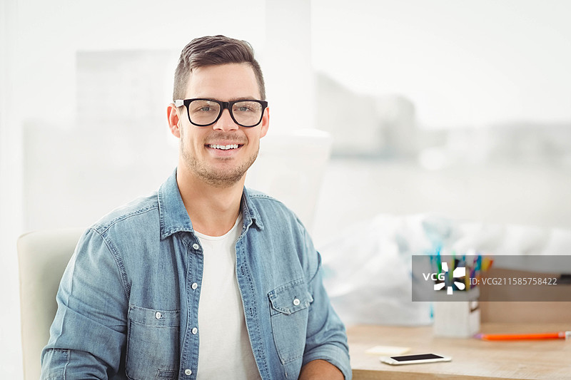 微笑的男人坐在桌子旁，手里拿着智能手机和彩色铅笔，靠近明亮的窗户。图片素材