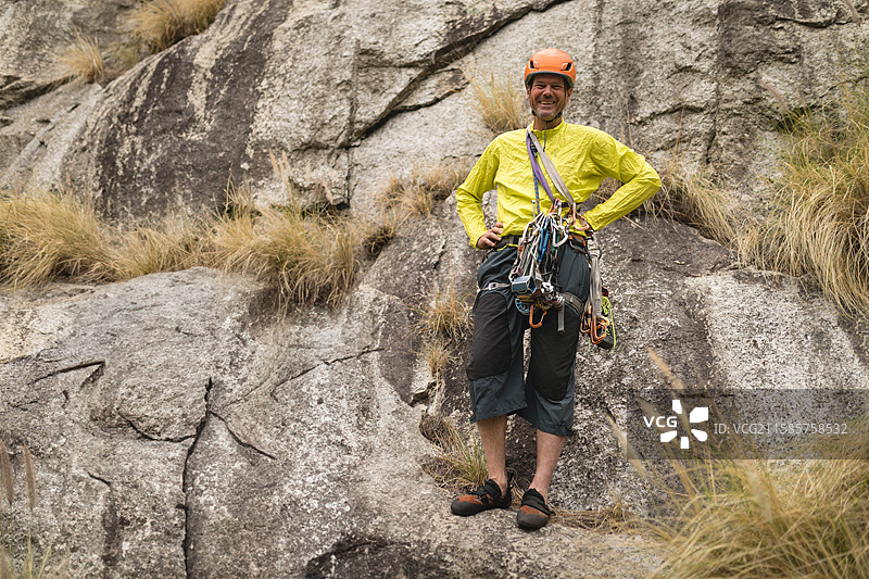 穿着黄色夹克和头盔，系着安全带和绳索的老年男性攀岩者站在岩石悬崖上图片素材