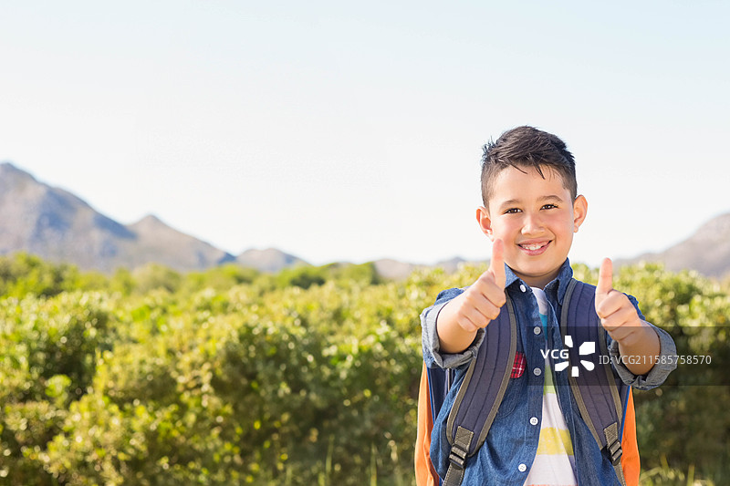 男孩在乡村户外站着，竖起大拇指，穿着牛仔衬衫和背包，留有空白空间。图片素材