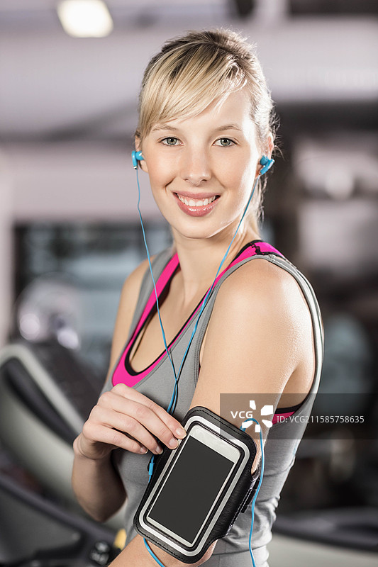 女性在健身房的有氧运动器械上佩戴耳机，调整上臂的智能手机臂带图片素材