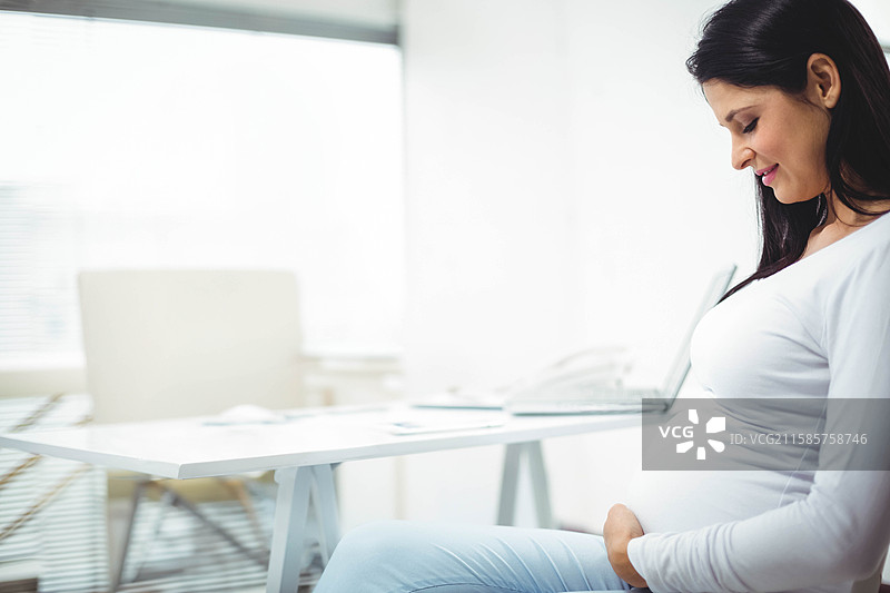 孕妇坐在现代办公室的桌子旁，手托着肚子，旁边有笔记本电脑和手机，留有空白空间。图片素材