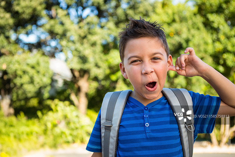 男孩在公园里背着背包，拉着耳朵做出顽皮的表情，留有空白空间。图片素材