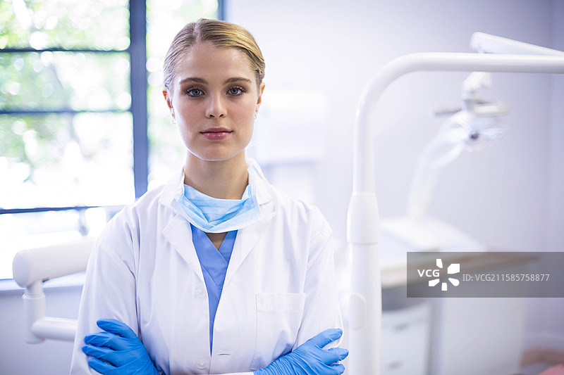 女牙医双臂交叉站在牙科诊所的窗边，旁边有吊灯。图片素材