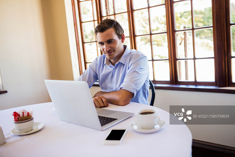 穿着扣领衬衫的男子在明亮的房间里，坐在圆形白色桌子旁用笔记本电脑打字，旁边放着一部智能手机。图片素材