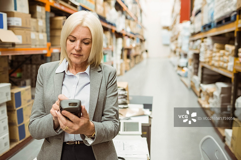 穿着西装的老年女性在仓库过道中手持扫描仪扫描箱子图片素材