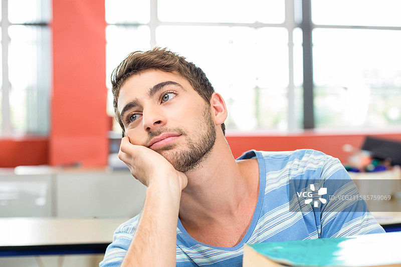 男学生坐在学校的课桌前，仰望着红色柱子旁的大窗户，桌上放着教科书。图片素材