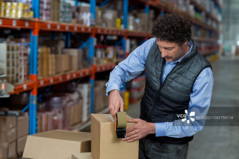 男性工人在仓库中使用胶带分配器封箱，旁边是货架，留有空白空间。图片素材