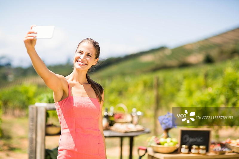 穿着裙子的女人在葡萄园摊位前站着，手持智能手机自拍，旁边有篮子，留有空白空间。图片素材