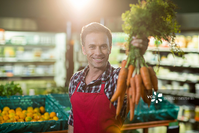 穿着红色围裙的男性杂货店员工在生鲜区向镜头展示一把胡萝卜图片素材