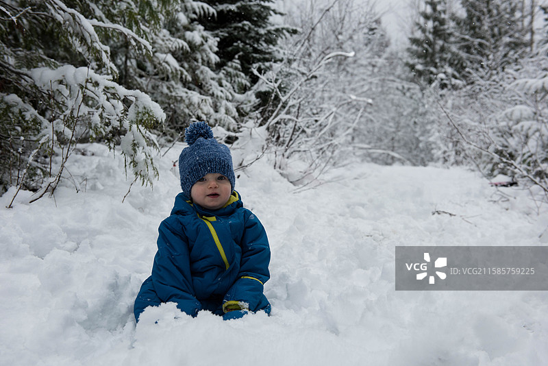 穿着蓝色冬季雪服和针织帽的小男孩坐在森林小径上的厚雪中图片素材
