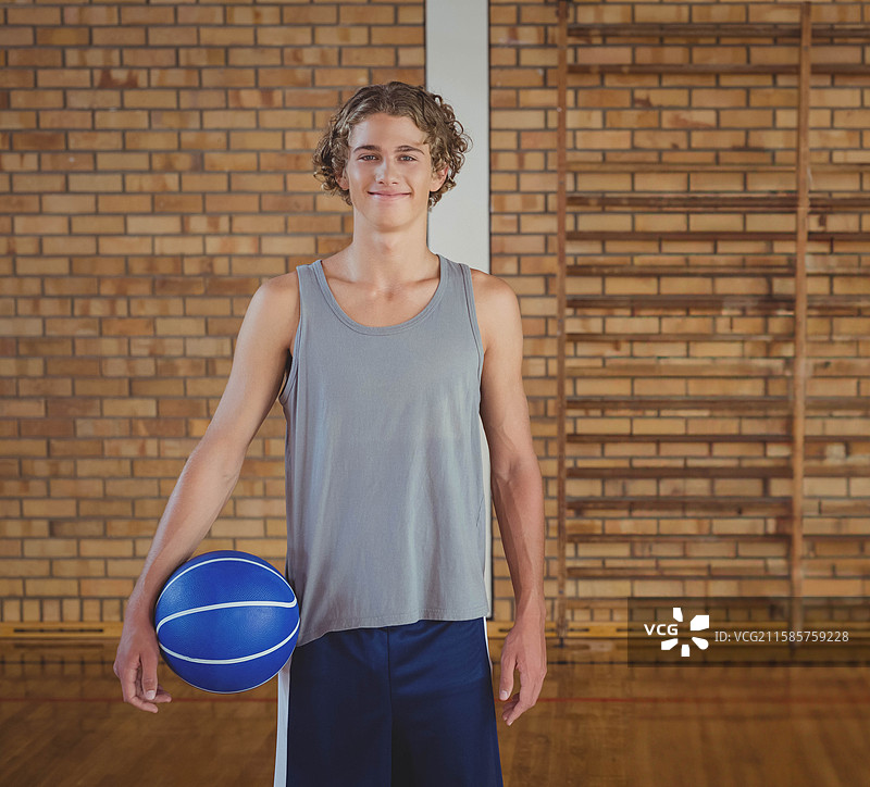 微笑的少年运动员在学校体育馆的砖墙前手持蓝色篮球图片素材