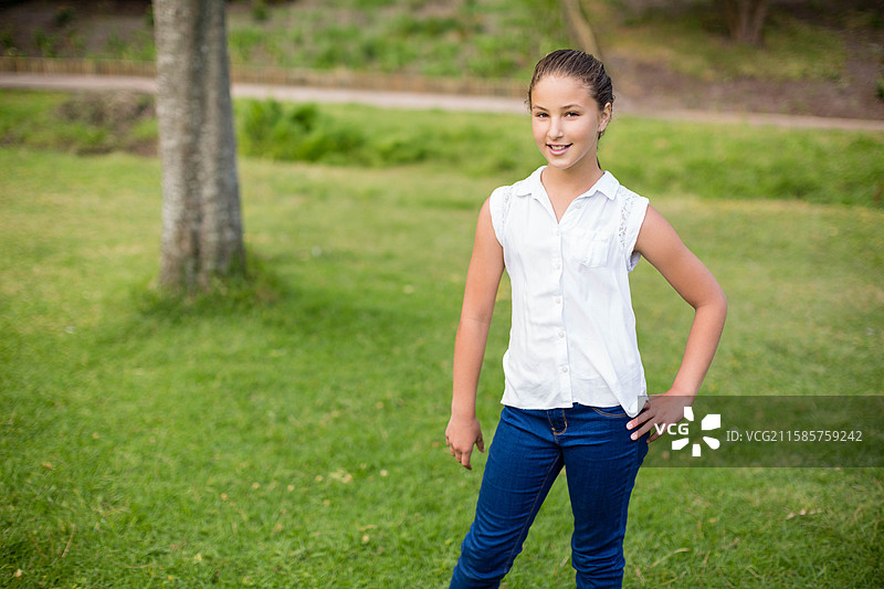 公园里一名女孩站在草坪上，旁边是一棵成熟树干和一条泥土小路。图片素材