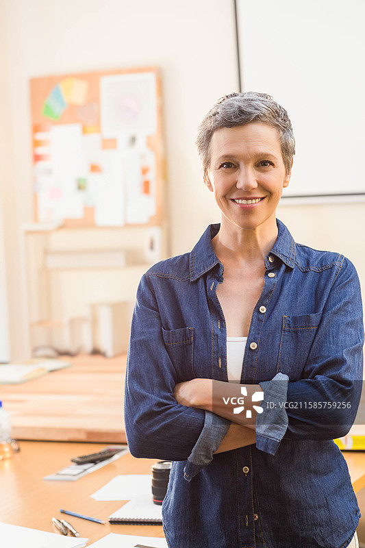 一位年长女性站在工作室里，双臂交叉，旁边有桌子、相机镜头和公告板。图片素材