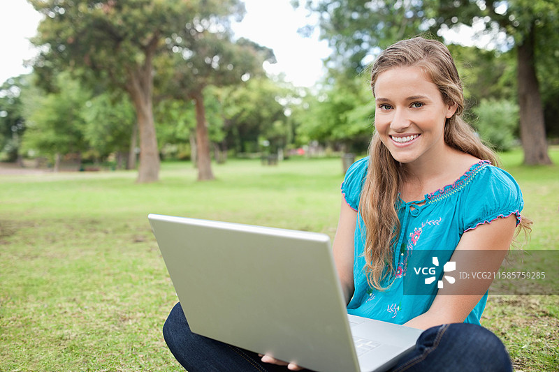 在公园的草坪上，盘腿坐着的女性正在使用银色笔记本电脑，留有空白空间。图片素材