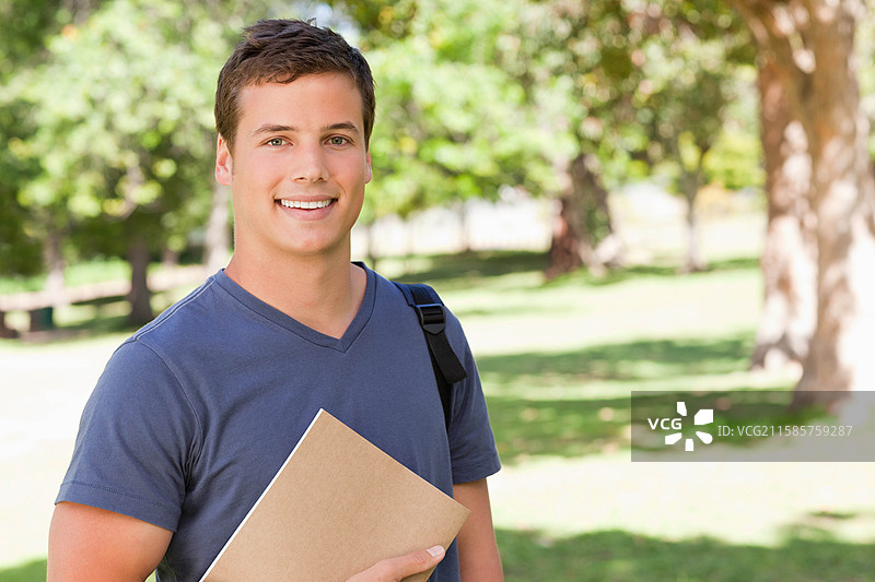 男学生穿着蓝色T恤，在校园草坪上，阳光下手持文件夹和背包。图片素材