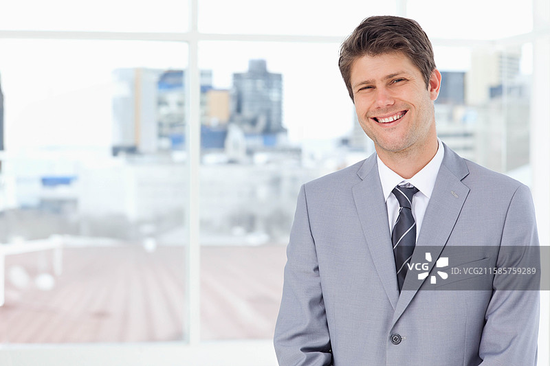 穿着灰色西装的中年男子在办公室窗前微笑，窗外是城市天际线，留有空白空间。图片素材
