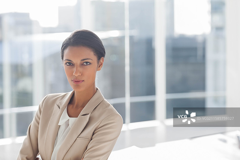女商人在办公室窗前双臂交叉，俯瞰模糊的天际线，旁边是一张白色办公桌。图片素材