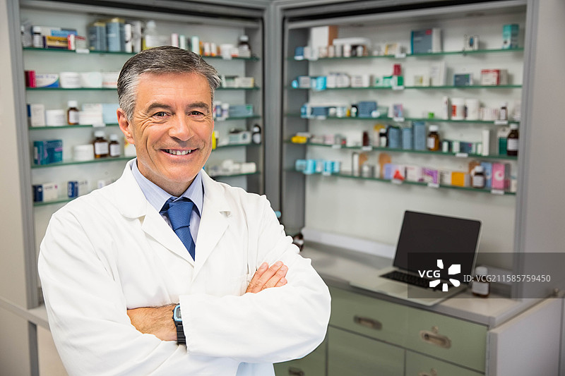 穿着实验室外套的老年男性药剂师站在柜台前，旁边有笔记本电脑和药瓶，留有空白空间。图片素材