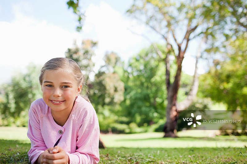 穿着粉色开衫和紫色上衣的女孩躺在公园的草坪上，留有空白空间。图片素材