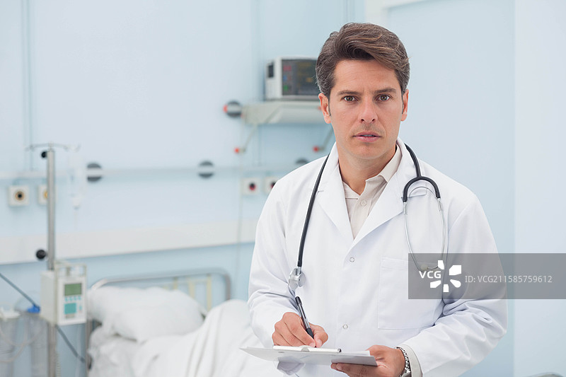 穿着实验室外套的男性医生手持夹板和听诊器，正在医院检查生命体征监测仪。图片素材