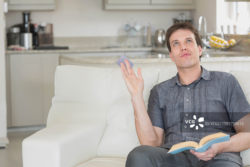 中年男子在休闲厨房的白色沙发上阅读平装书，并将智能手机扔在一旁，留有空白空间。图片素材