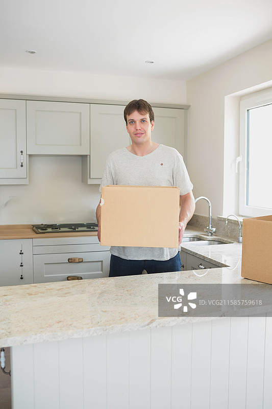 中年男子在大理石家居厨房岛后面拿着纸箱，窗外自然光照射进来。图片素材