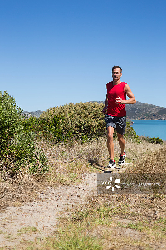 穿着红色衬衫和跑鞋的男子在宁静的海湾附近的泥土小径上奔跑，留有空白空间。图片素材